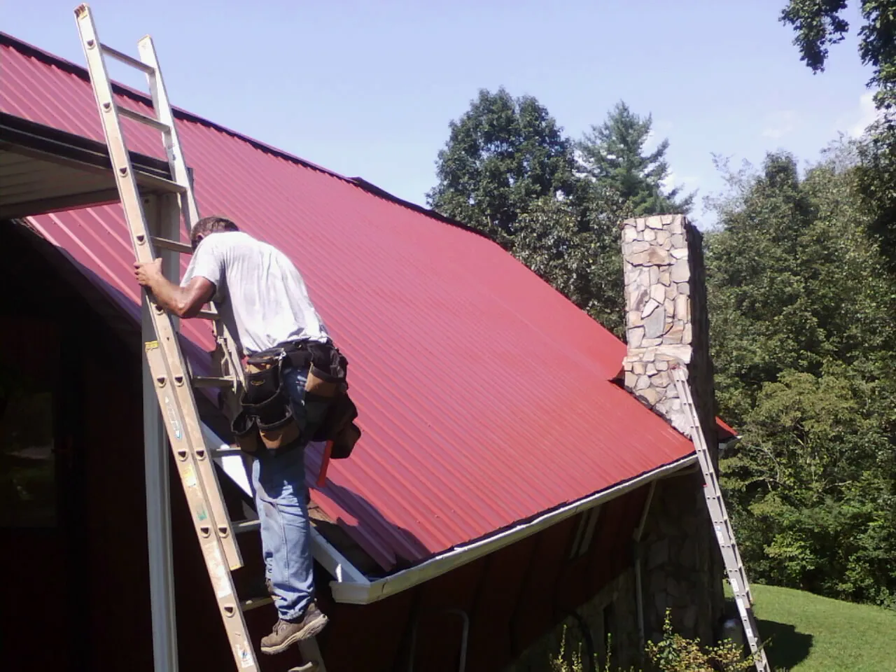 Roofer on a ladder beside a red metal roof and stone chimney — Soffit & Fascia Repair by Roofing Brothers in Sterling Forest
